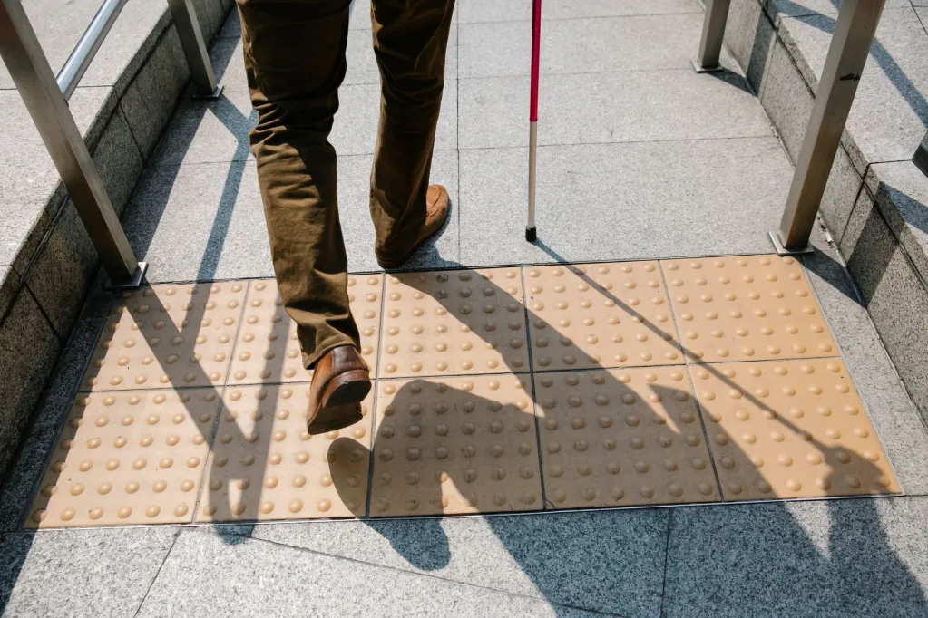 A person with a white cane walking on textured tactile paving with a shadow visible on the ground.